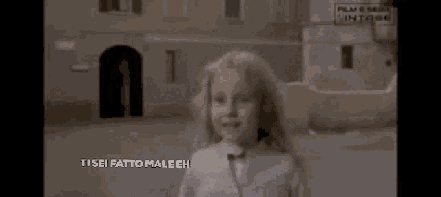 a little girl is standing in front of a building with the words ti sei fatto male eh on the bottom