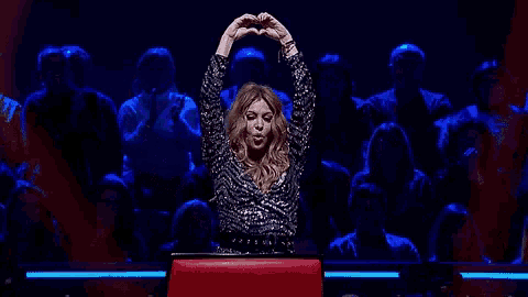 a woman is making a heart shape with her hands while sitting in front of a crowd .