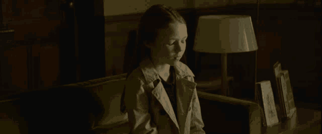 a girl in a trench coat sits on a green couch in a dark room