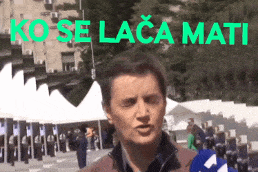 a man in a brown jacket is talking in front of a sign that says ko se laca mati