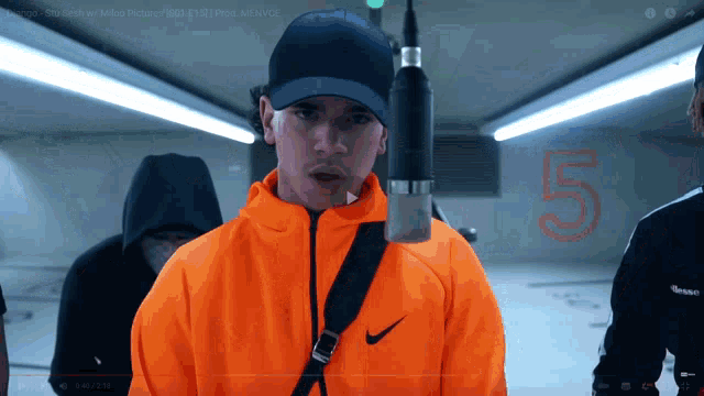 a man in an orange nike jacket stands in front of a microphone and the number 5