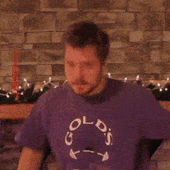 a man in a purple shirt is standing in front of a fireplace with christmas lights on it .
