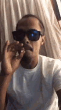 a man wearing sunglasses is smoking a cigarette in front of a white curtain .
