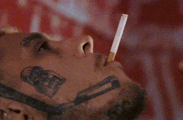 a man with a tattoo on his face is smoking a cigarette with a needle in his mouth .