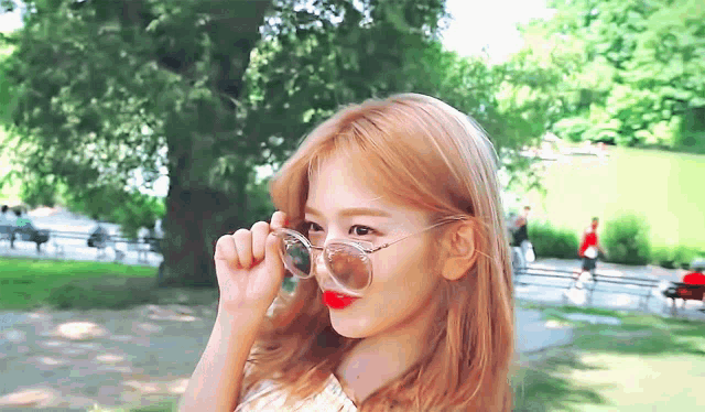 a woman wearing sunglasses and red lipstick looks at the camera in a park