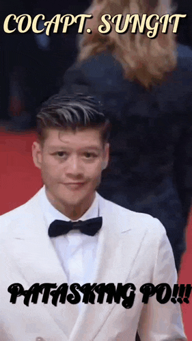 a man in a white suit with a black bow tie is on a red carpet
