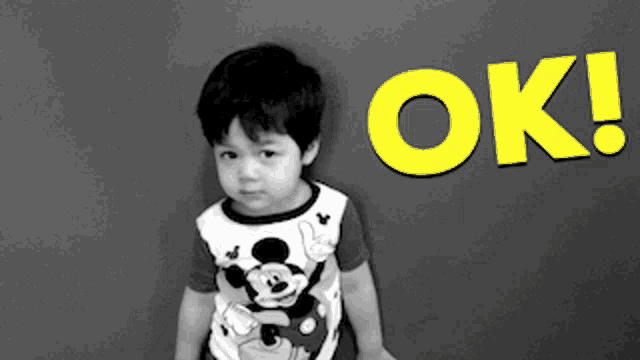 a young boy wearing a mickey mouse shirt is standing in front of a wall that says ok