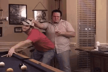two men are playing pool in a living room and one of them is wearing a hat