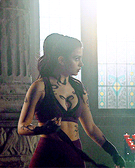 a woman with tattoos on her arms and chest is standing in front of a window holding a sword .