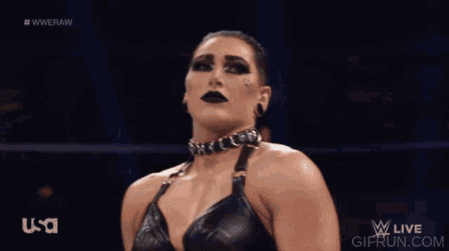 a female wrestler wearing black makeup and a choker stands in front of a screen that says usa