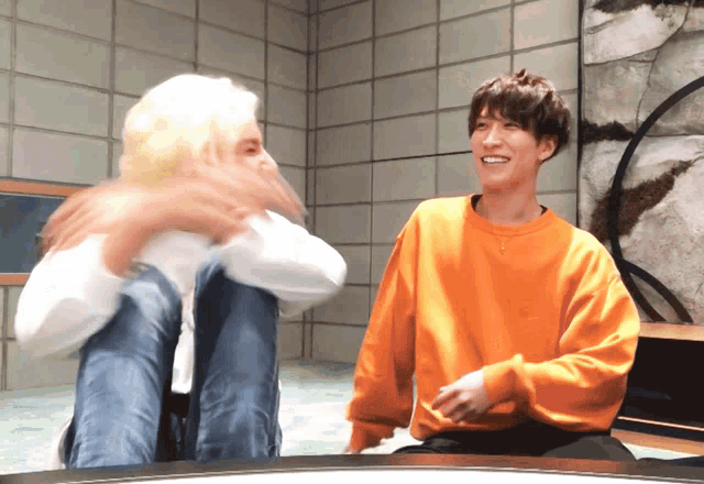 a man in an orange sweatshirt sits next to another man in a white shirt
