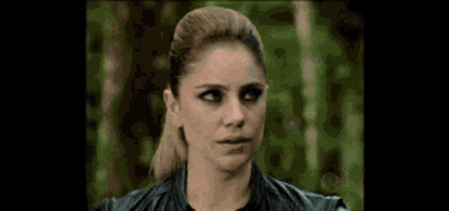 a woman in a leather jacket is standing in the woods looking at the camera .