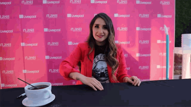 a woman in a red jacket sits at a table in front of a wall that says liverpool