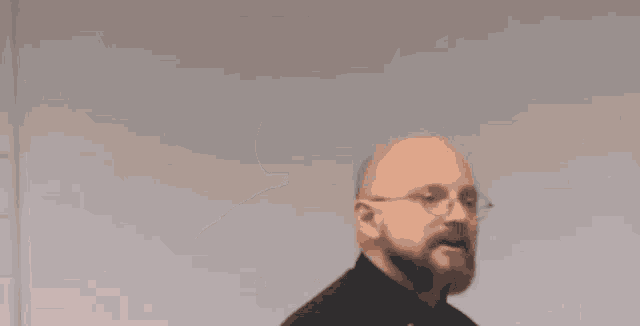 a bald man with a beard is wearing a black chef 's jacket with a fork on his sleeve .