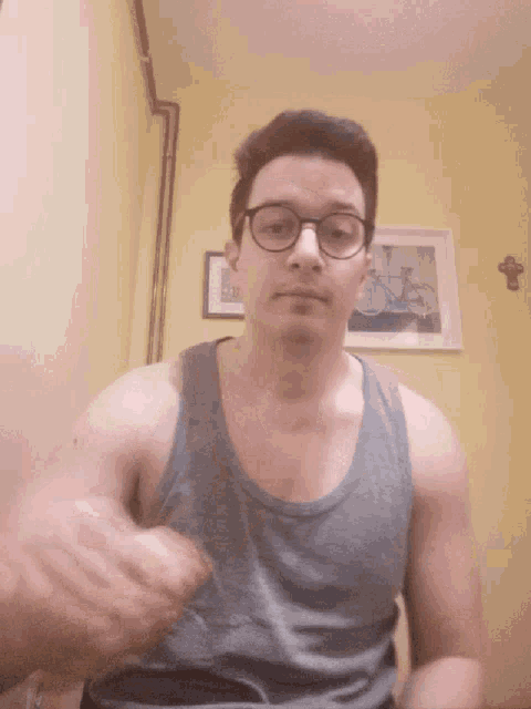 a man wearing glasses and a grey tank top is sitting in front of a yellow wall