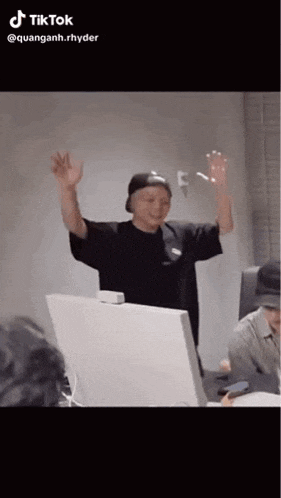 a man in a black hat is standing in front of a computer with his arms in the air .