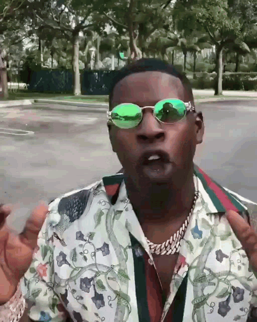 a man wearing green sunglasses and a floral shirt stands in a parking lot