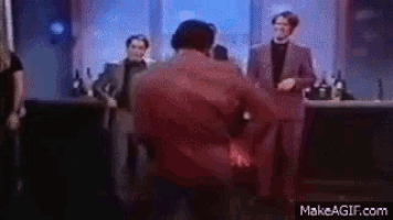 a man in a red shirt is dancing in front of a group of men in suits in a bar .