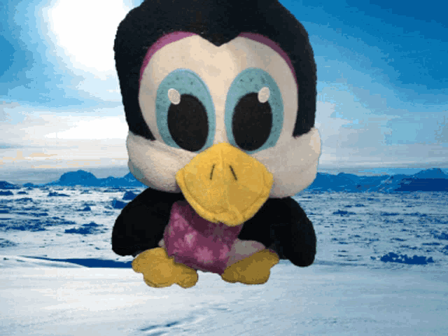 a stuffed penguin with a yellow beak is sitting in the snow with mountains in the background