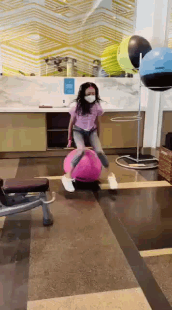 a young girl wearing a mask is jumping on a pink exercise ball .