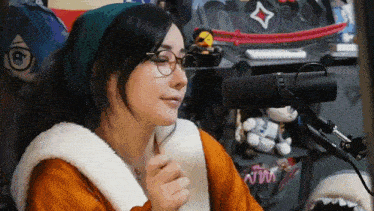 a woman wearing glasses and a beanie is sitting in front of a microphone with a stuffed animal in the background