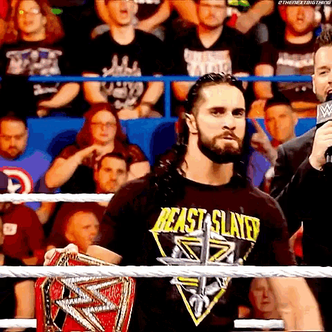 a man in a beast slayer shirt is standing in a wrestling ring holding a championship belt .