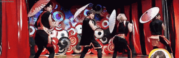 a group of people are dancing on a stage with umbrellas and fans
