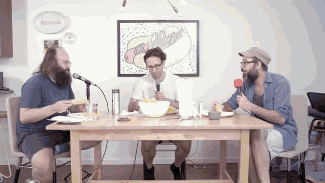 three men are sitting at a table with a picture of a hot dog behind them