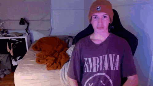 a young man wearing a nirvana shirt and a beanie stands in front of a bed