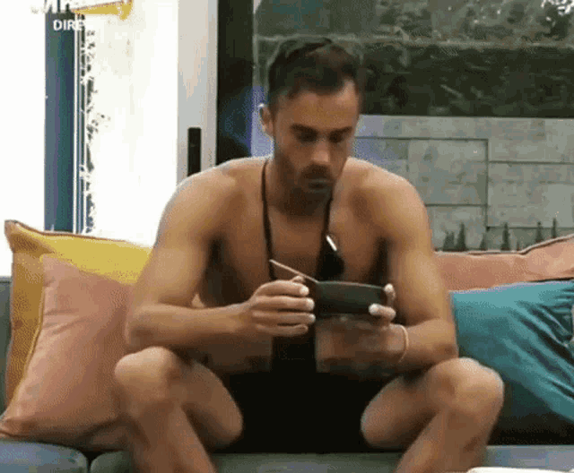 a shirtless man is sitting on a couch looking at a cell phone