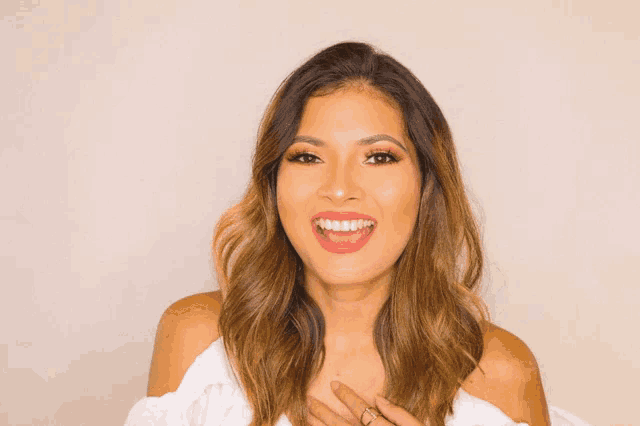 a woman in a white off the shoulder top smiles with her hand on her chest