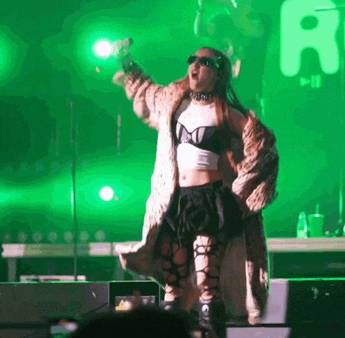 a woman in a fur coat is singing into a microphone in front of a green background with the letter a on it
