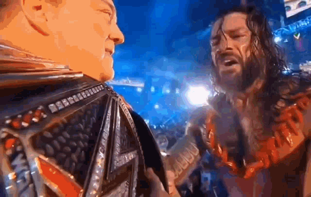 two wrestlers are standing next to each other on a stage . one of the men is wearing a championship belt .