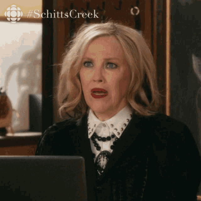 a woman sitting in front of a laptop with a #schitts creek logo behind her