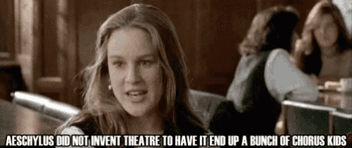 a woman is sitting at a table with a bunch of chorus kids in the background .