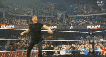 a man in a black shirt is standing in a wrestling ring with a crowd of people watching .