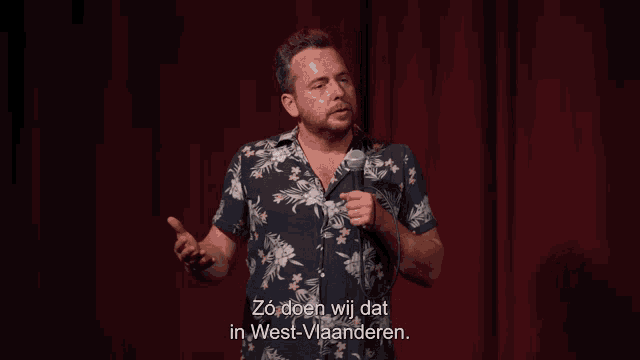 a man in a floral shirt stands on a stage holding a microphone and says zo doen wij dat in west-vlaanderen