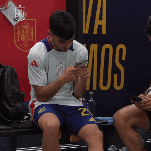 a man sitting on a bench looking at his phone in front of a sign that says " va mos "