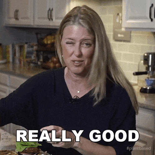a woman in a kitchen says " really good " while holding a hamburger