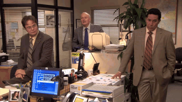 three men in suits are standing in front of a computer screen that says dunder mifflin