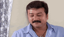 a man with a mustache wearing a blue shirt and a necklace is sitting in front of a window .