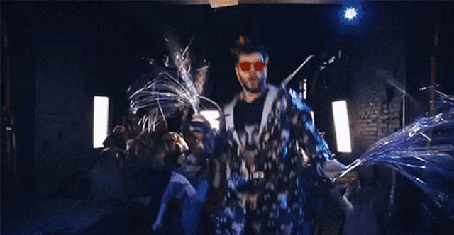 a man in a camouflage suit and red sunglasses is dancing in a dark room in front of a crowd .