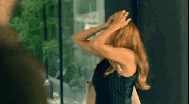 a woman in a black dress adjusts her hair while standing in front of a window