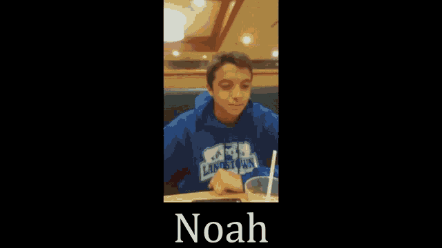 a boy in a blue hoodie is sitting at a table drinking from a cup with the name noah written on the bottom
