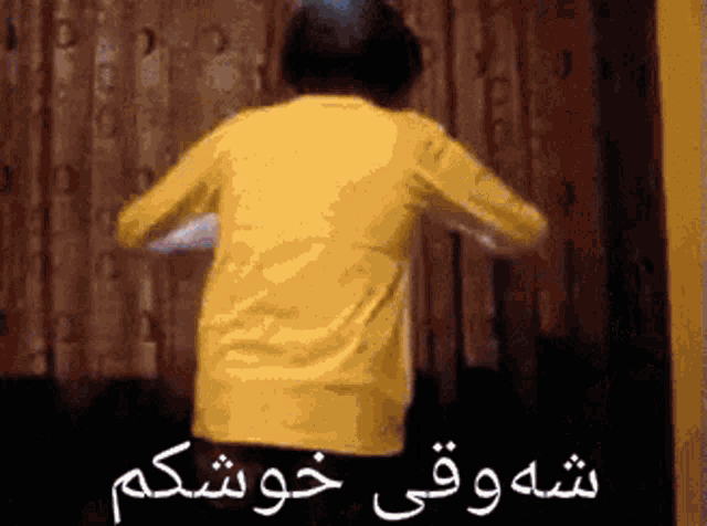 a man in a yellow shirt is standing in front of a wooden wall with arabic writing on it .