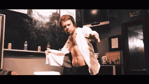 a man without a shirt is dancing in a room with a tv and bottles on a shelf .
