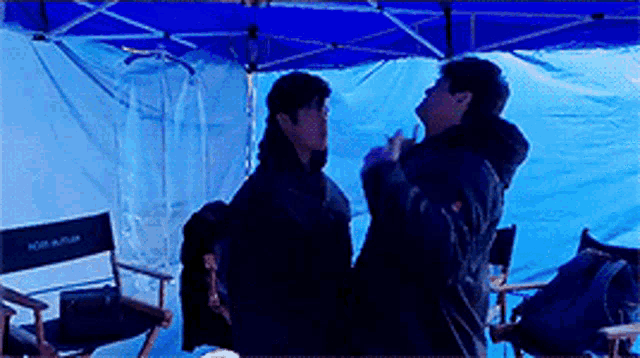 two people are standing under a blue tent with a director 's chair in the background