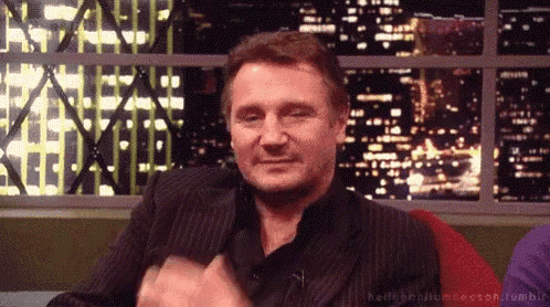 a man in a suit is sitting in front of a window and making a funny face .