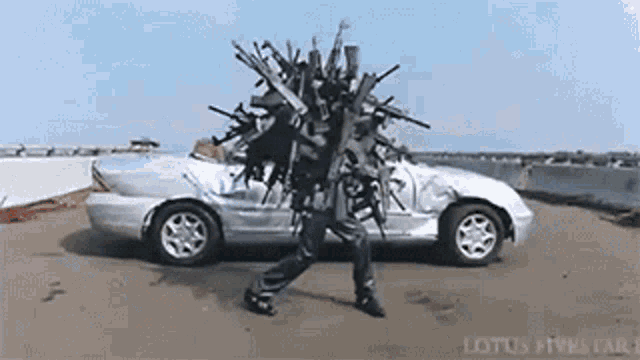 a man is carrying a bunch of guns on his head next to a silver car
