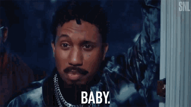 a man with long hair and a beard is standing in front of a window and says baby .